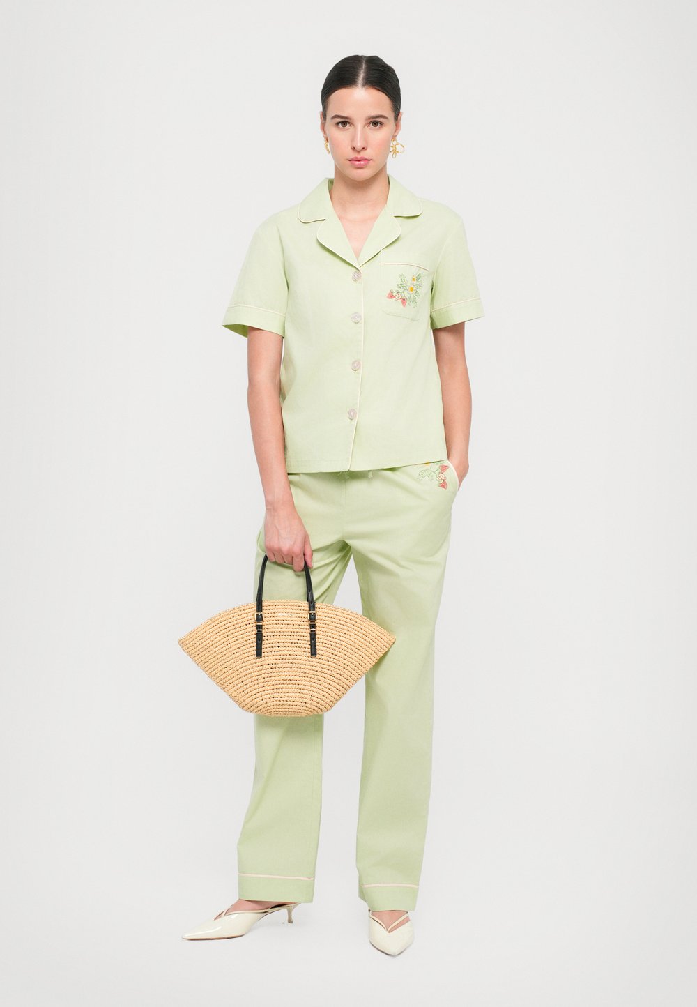 Haut à manches courtes, boutonné, vert clair avec broderies florales, associé à un pantalon assorti. Tient un sac en paille texturé avec des poignées noires.