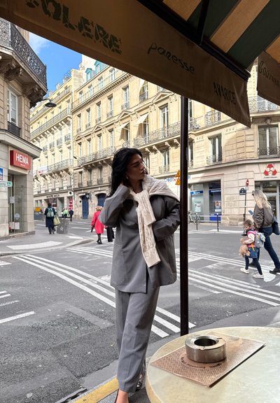 Chaqueta y pantalones de traje gris, combinados con una bufanda de punto color crema. Vista de un café al aire libre con la calle y peatones de fondo. Cenicero de metal sobre la mesa.