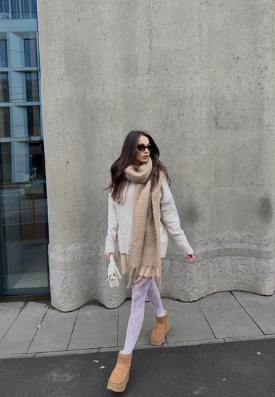 Suéter oversized beige claro, bufanda beige, medias blancas con estampado, botas cortas color marrón claro con suelas gruesas, sosteniendo un pequeño bolso blanco, de pie al aire libre.