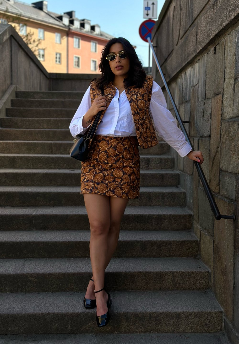 Weste mit braunem Blumenmuster und passendem Miniröckchen, kombiniert mit einem weißen Hemd und schwarzen High-Heels; steht auf Steinstufen.