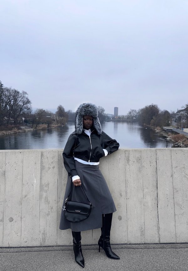 Kobieta w czarnej kurtce, szarej spódnicy, wysokich czarnych kozakach i futrzanej czapce stoi na betonowym moście nad rzeką pod zachmurzonym niebem.