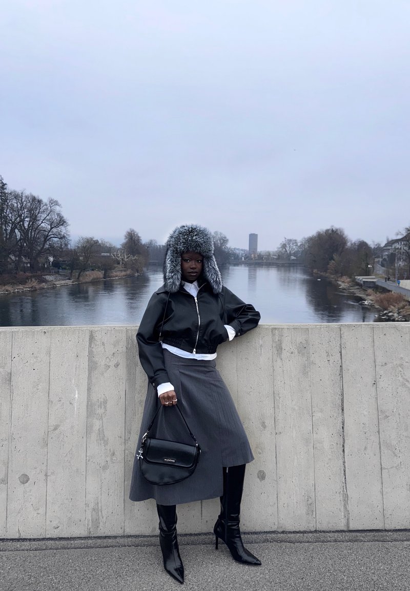 Femme en veste noire, jupe grise, bottes hautes noires et chapeau en fourrure debout sur un pont en béton au bord de la rivière sous un ciel couvert.