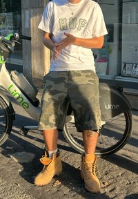 Hnedé šnurovacie topánky, kamuflážové šortky a biele tričko so zeleným nápisom. Vidieť bicykel s zeleným logom "Lime".