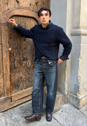 Suéter de punto grueso azul marino, jeans sueltos azul oscuro y botines de cuero marrón. De pie contra una puerta rústica de madera con vetas y desgaste visibles.