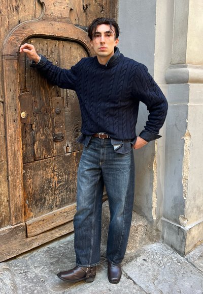 Suéter de punto grueso azul marino, jeans sueltos azul oscuro y botines de cuero marrón. De pie contra una puerta rústica de madera con vetas y desgaste visibles.