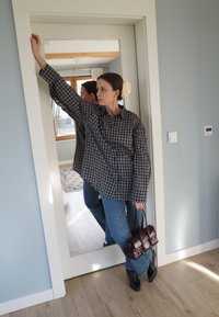 Chemise oversize grise à carreaux, jean bleu évasé, bottines noires pointues, et petit sac à main violet foncé. En arrière-plan, un miroir et un sol en bois clair.