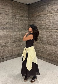 Brun ærmeløs top og lang brun plisseret nederdel, bundet gul stof ved taljen, brune sko med mønstret design, stående i hjørnet.