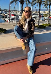 Manteau imprimé léopard, haut blanc, jeans bleu clair et chaussures en suède beige. Assis sur un rebord en pierre avec une marina en arrière-plan.