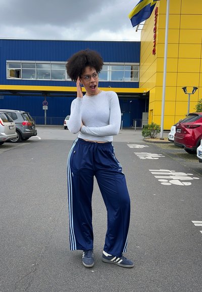 Una persona lleva una camiseta de manga larga gris claro y pantalones deportivas azul marino con rayas blancas en los lados. Está de pie al aire libre cerca de un aparcamiento.