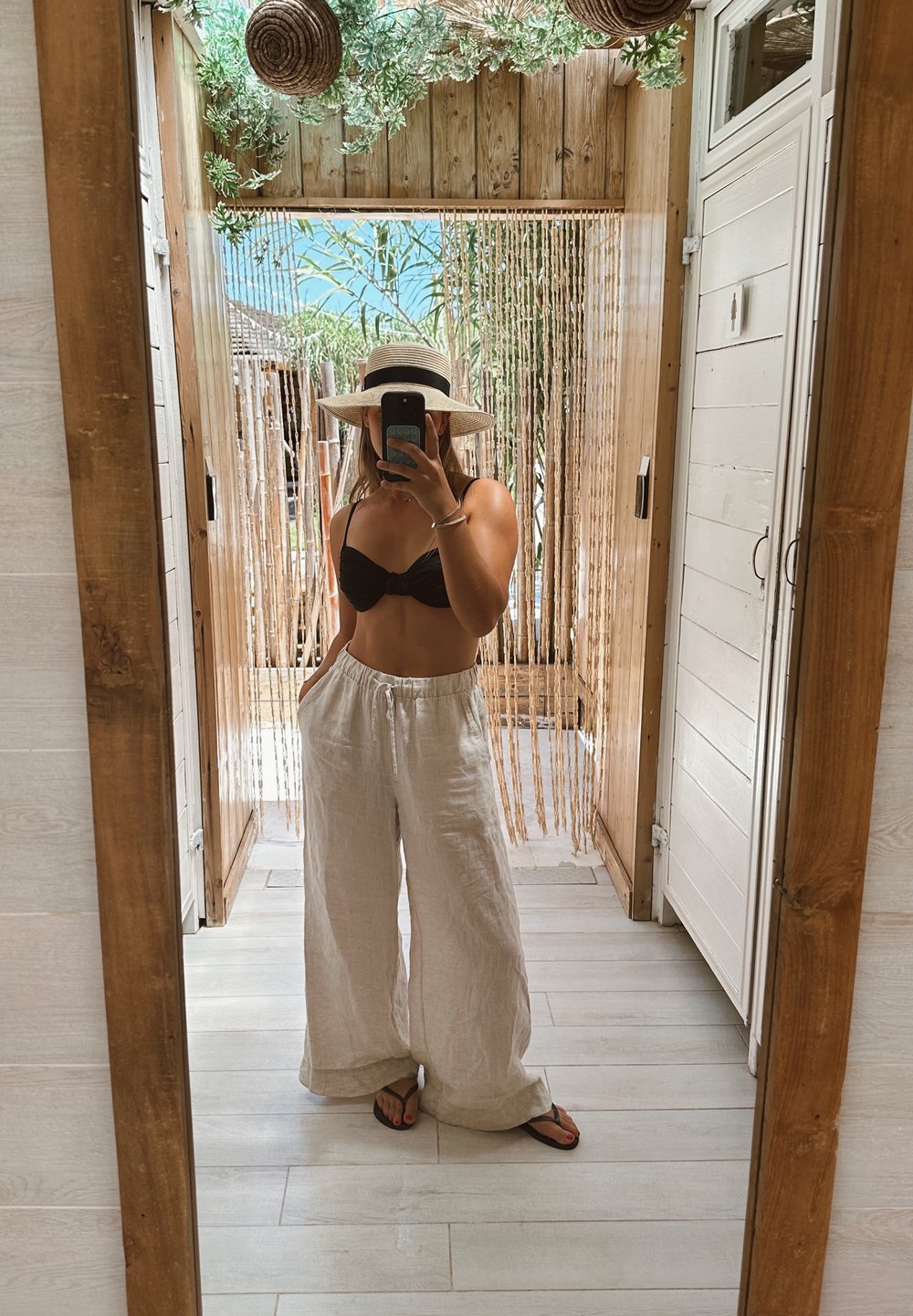 Linnen witte wijde broek, zwarte bikini top en een strohoed. Staande in een houten interieur met een spiegel en natuurlijke decoratie.