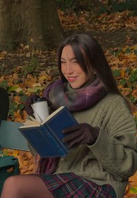 Grønn kabelstrikket genser, rutet skjørt og brune hansker. Holder en blå bok med gullakenter og en hvit kaffekopp i en park med høstløv.