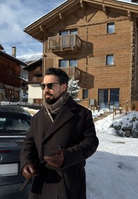 Manteau en laine foncé avec de gros boutons et une ceinture, gants en cuir marron et écharpe à motifs. Paysage enneigé et chalet en bois en arrière-plan.