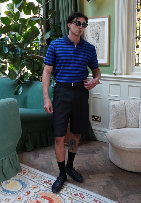 Homme portant des lunettes de soleil, un polo bleu à rayures, un short noir et des chaussures noires, debout dans une pièce décorée avec des rideaux verts et des œuvres d'art encadrées.