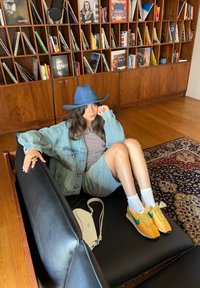 Jeansjakke og skjørt, stripete topp, blå denimhatt, gule og grønne sneakers, svart lærsofa, og hvit håndveske på tregulv.