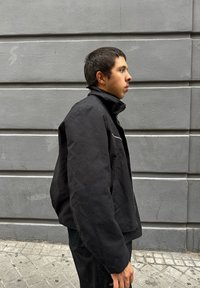 Giovane con capelli corti scuri e giacca nera in piedi di lato contro un muro di cemento grigio all'aperto.