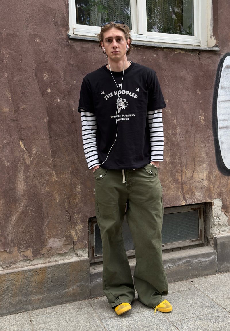 Jeune personne avec des manches longues rayées sous un t-shirt graphique noir, pantalon vert ample, chaussures jaunes, debout contre un mur marron usé avec une fenêtre.