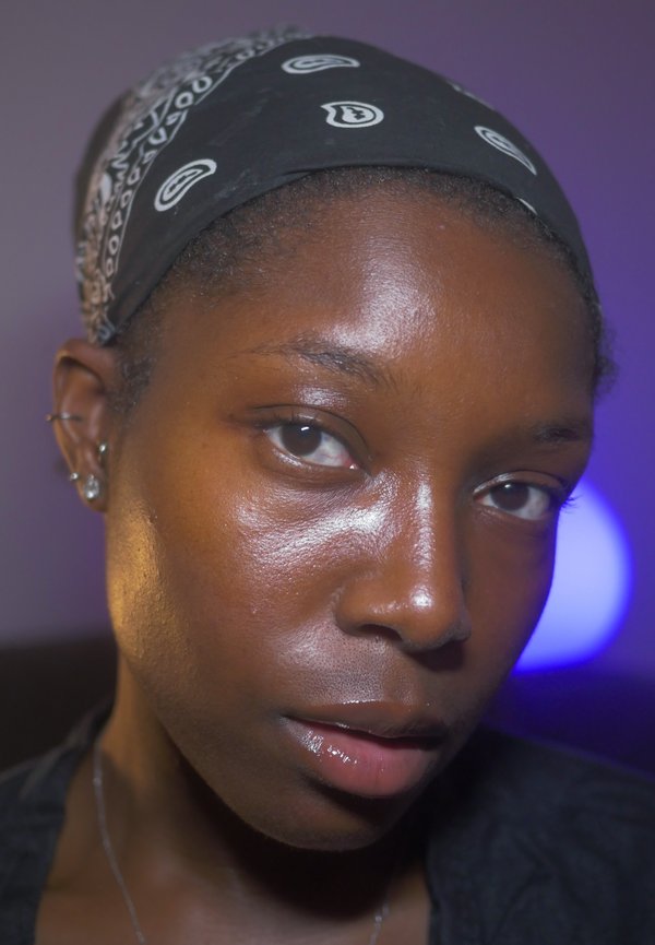 Gros plan d'une personne à la peau foncée portant un bandana noir avec des motifs blancs et plusieurs piercings à l'oreille sur un fond violet.