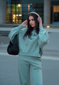Mintgrønn Nike genser og joggebukser, med en stor logo i svart, paret med beige hodetelefoner og en svart veske.