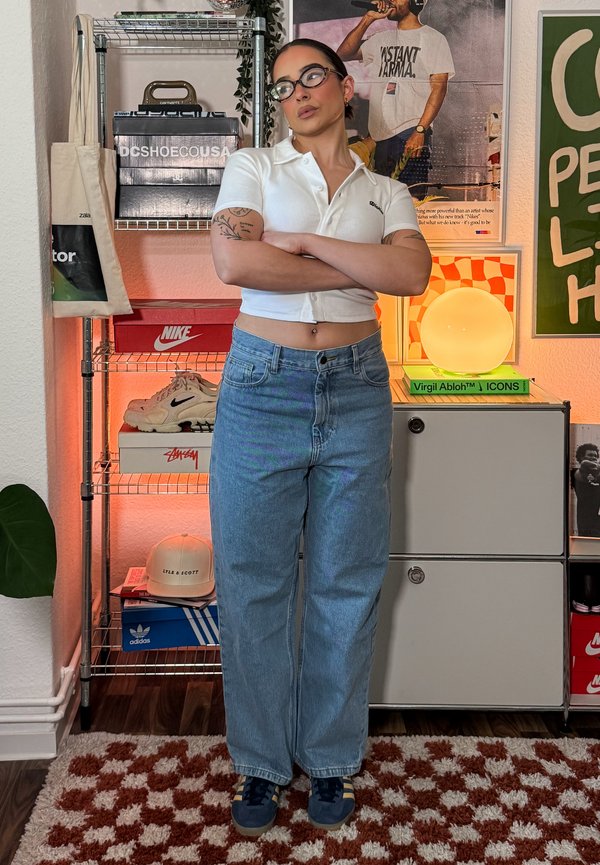 Tânăr cu ochelari, tatuaje, tricou alb scurt și blugi albaștri stând cu brațele încrucișate într-o cameră cu pantofi sport și postere.