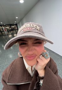 Brun keps med "BROOKLYN 1947" broderat i beige, bärs med en brun jacka. Stort ring med blå detaljer och silverfärgade beslag.