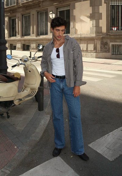 Camisa de cuadros en marrón y blanco, camiseta sin mangas blanca, jeans de denim azul, zapatos oscuros; de pie junto a un scooter crema en un entorno urbano.