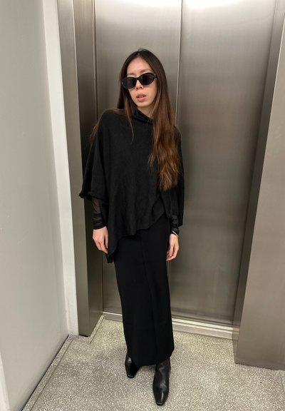 Top tipo poncho negro hecho de tejido suave, superpuesto a una falda negra ajustada. Los accesorios incluyen gafas de sol oscuras y botas negras puntiagudas.