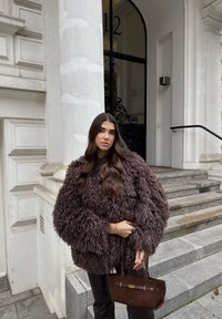 Jachetă maro pufoasă cu mâneci lungi și largi, însoțită de o geantă mică, maro închis, cu un accent auriu. Trepte de piatră în fundal.