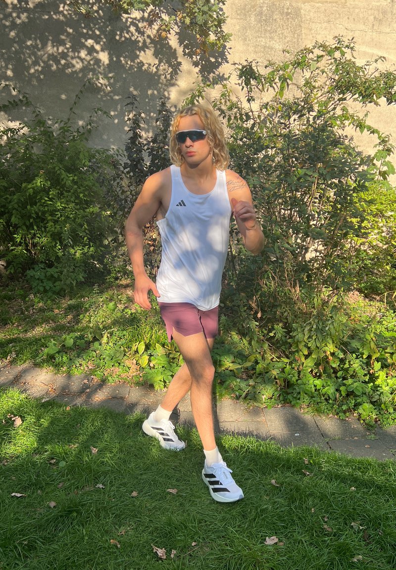 Wit mouwloos sportshirt met zwart logo, paarse shorts en witte sneakers met zwarte accenten. Weelderige groene achtergrond met schaduwen.