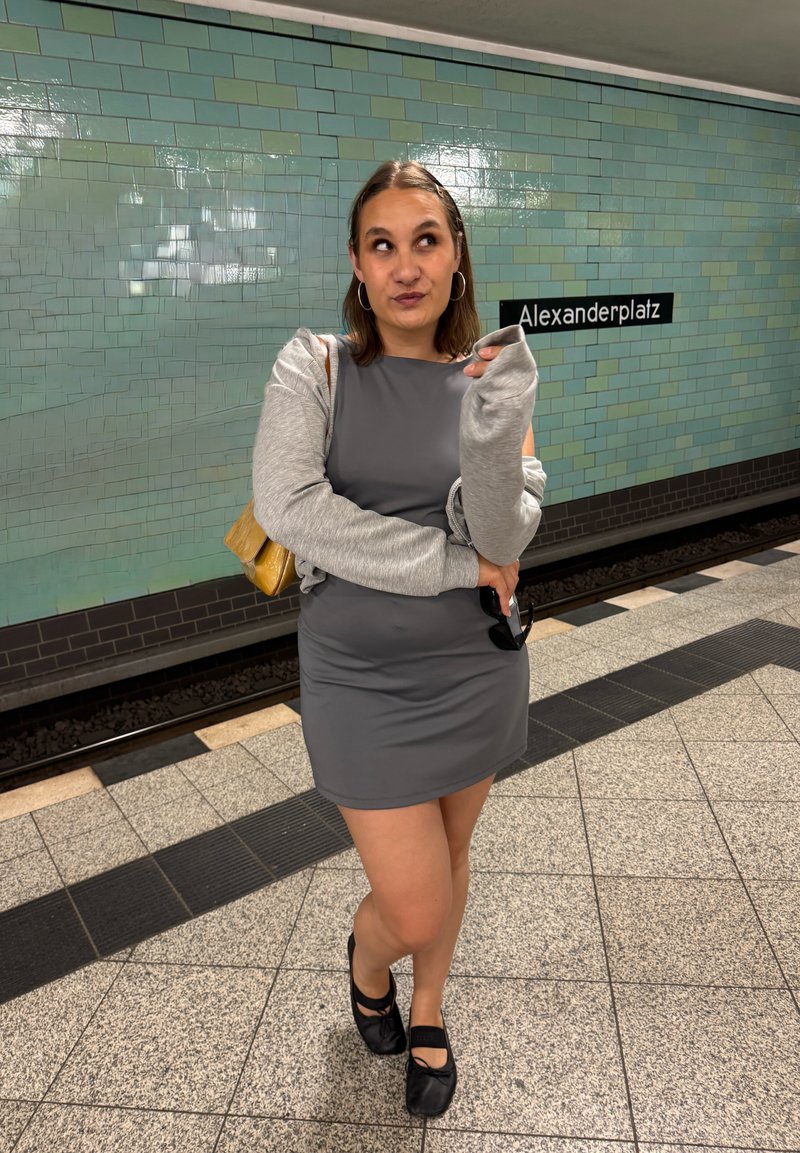 Woman in gray dress and sweater stands on Alexanderplatz subway platform, looking up with a thoughtful expression.