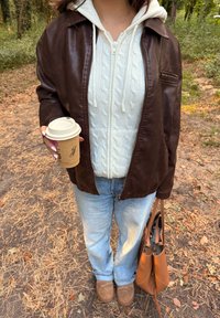 Veste en cuir marron par-dessus un sweat à capuche en tricot blanc, jeans bleu clair, bottes marron, tenant une tasse de café et portant un sac à main beige.