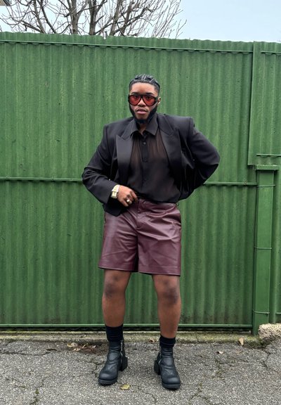 Blazer negro sobre una blusa negra de tela transparente, pantalones cortos de cuero burdeos y botas negras de tobillo. Grandes gafas de sol rojas y un reloj de pulsera plateado complementan el look.