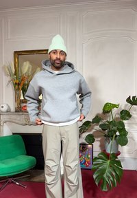Sweat à capuche gris avec poche avant et capuche à cordon, associé à un pantalon beige. Bonnet vert menthe clair. Intérieur avec des plantes et des œuvres d'art.