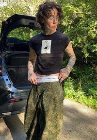Juodas trumpas marškinėliai su nuotraukų spauda, derinami su laisvomis, tekstūruotomis žaliomis kamufliažo kelnėmis. Matomas baltas Calvin Klein juostos kraštas.