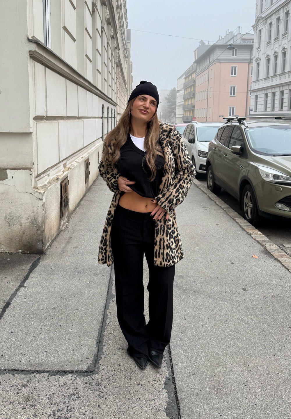 Leopardmönstrad kappa, svart topp och svarta vida byxor. Modellen bär en svart mössa, har långt hår och ljus hy, står på en gata.