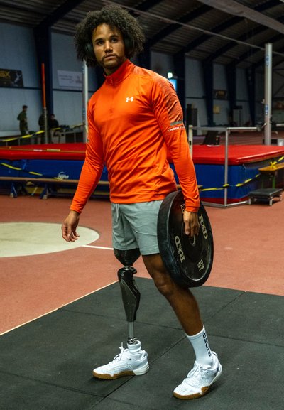 Atleta que lleva una camiseta de manga larga de color naranja brillante y pantalones cortos grises, sosteniendo un disco de pesas negro. Se destaca por su pierna protésica y zapatos deportivos blancos.