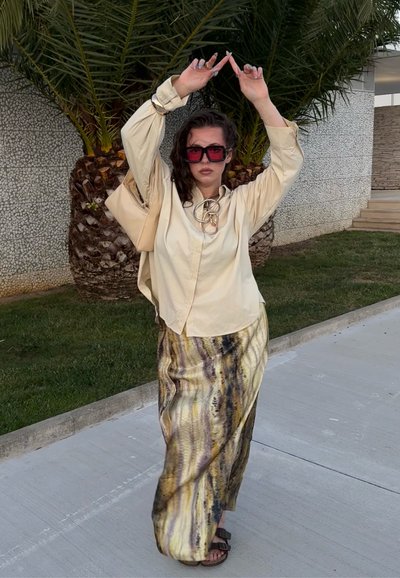 Camisa beige de botones con una falda texturizada que presenta patrones amarillos y marrones. Grandes gafas de sol rojas y un bolso beige completan el atuendo.