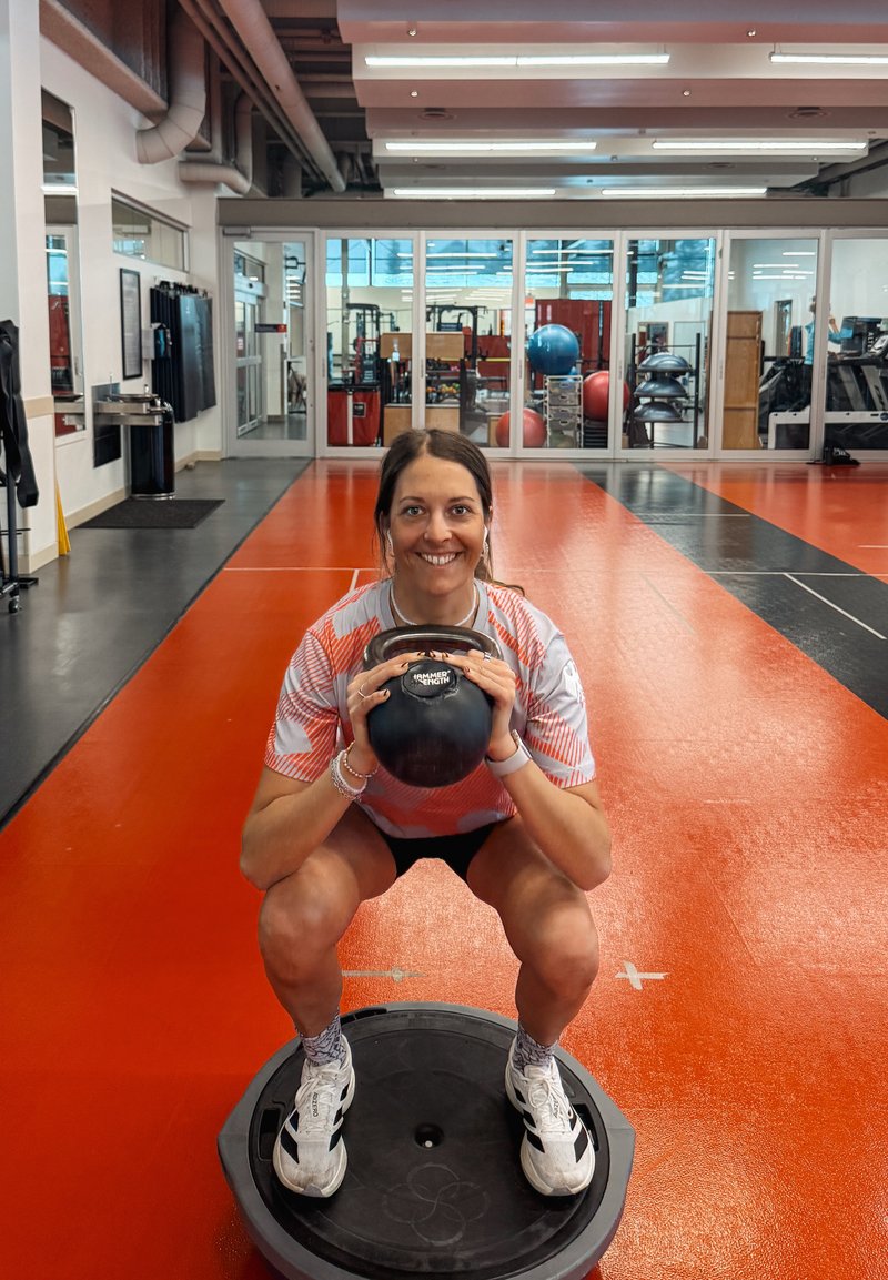 Γυναίκα κρατώντας kettlebell ενώ κάνει κάθισμα σε σανίδα ισορροπίας σε γυμναστήριο με κόκκινο και μαύρο δάπεδο και εξοπλισμό άσκησης στο βάθος.