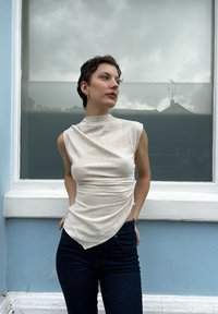 Μπέζ αμάνικο μπλουζάκι με ψηλό λαιμό και ζαρωμένο μπροστινό μέρος, σε συνδυασμό με σκούρο τζιν παντελόνι. Μινιμαλιστικός σχεδιασμός με διακριτική υφή.