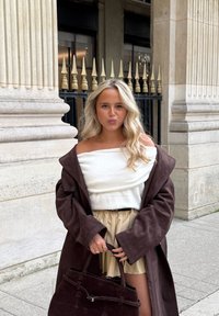 Brun semsket frakk, hvit skulderfri genser, beige plissert skjørt. Brun semsket håndveske med strukturert håndtak og sølvdetaljer.