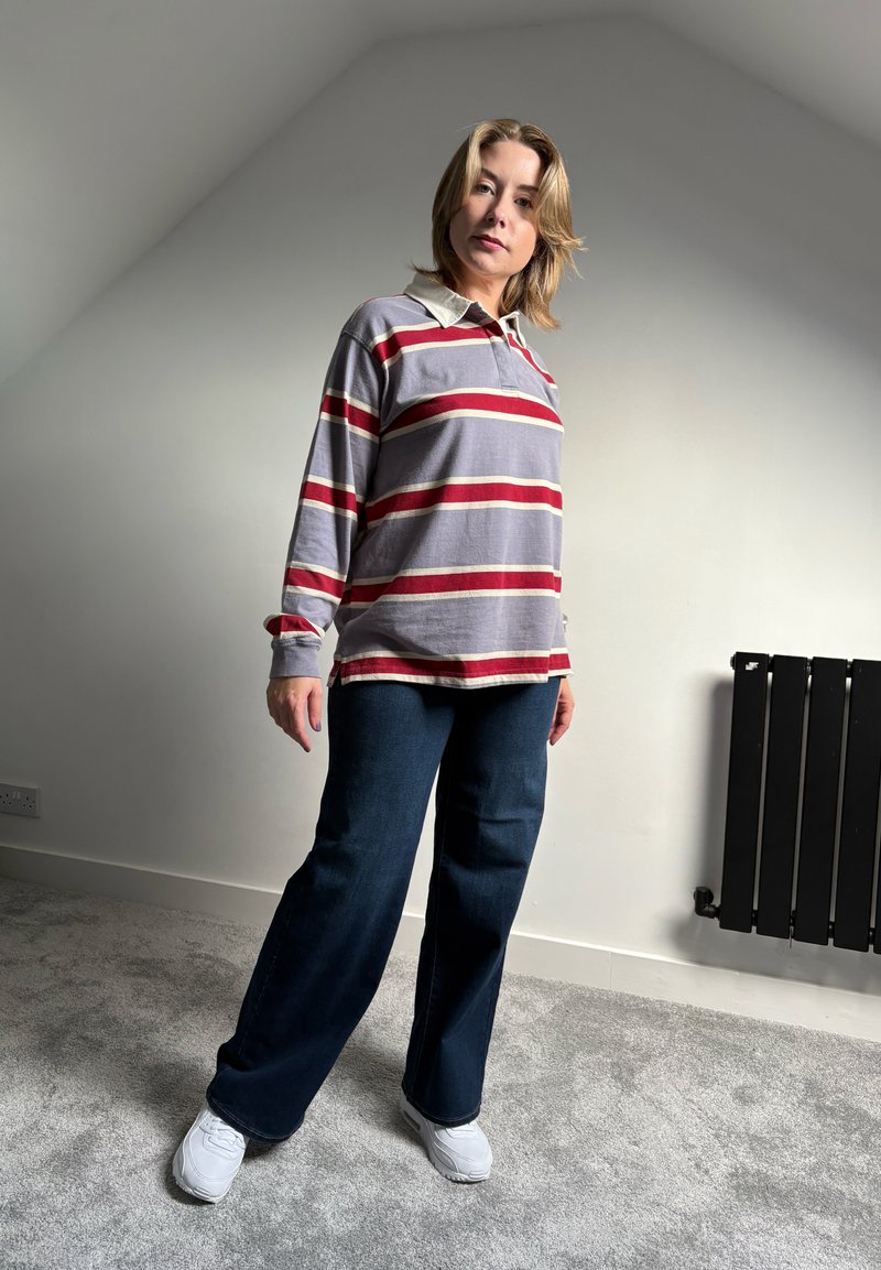 Langarm-Polohemd in Grau mit roten und weißen Streifen, kombiniert mit weit geschnittenen dunkelblauen Jeans und weißen Sneakers. Strukturierter Stoff, lässige Passform.