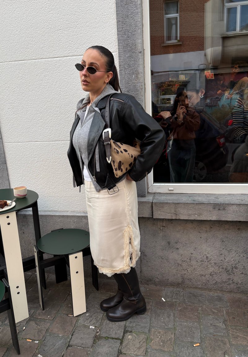 Femme portant une veste en cuir noire, un sweat à capuche gris, une jupe crème, des bottes noires et des lunettes de soleil, debout dans une rue pavée près d'une table de café avec café et gâteau.