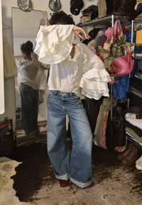 Fehér, fodros hosszú ujjú blúz világoskék, bő szárú farmerral párosítva. A háttérben ruhásállványok és egy tükör látható.