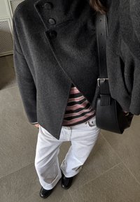 Persoană purtând o haină închisă la culoare, gri închis, cu nasturi, cămașă roz cu dungi negre, pantaloni albi, pantofi negri și având o geantă de umăr din piele neagră.