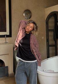 Model iført en sort skjorte under en rødternet overshirt og grå joggingbukser, stående nær et hvidt badekar og en struktureret væg.