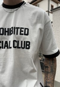 Hvit t-skjorte med svart tekst "PROHIBITED SOCIAL CLUB" i fet skrift. Kontrastfarge med svart ribbestrikket hals og ermekanter. Glatt bomullstekstur.