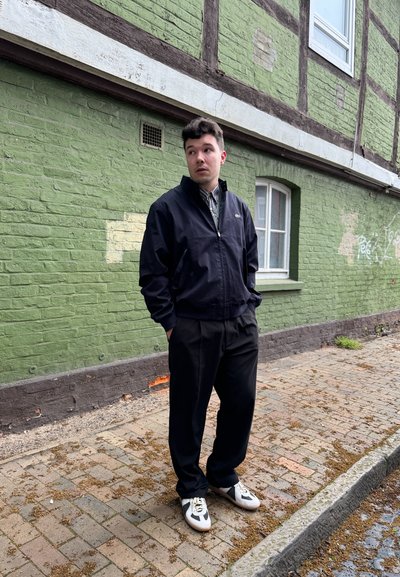 Chaqueta navy con cremallera, camisa de botones con estampado, pantalones negros y zapatillas blancas con detalles grises. De pie frente a una pared de ladrillos verdes.