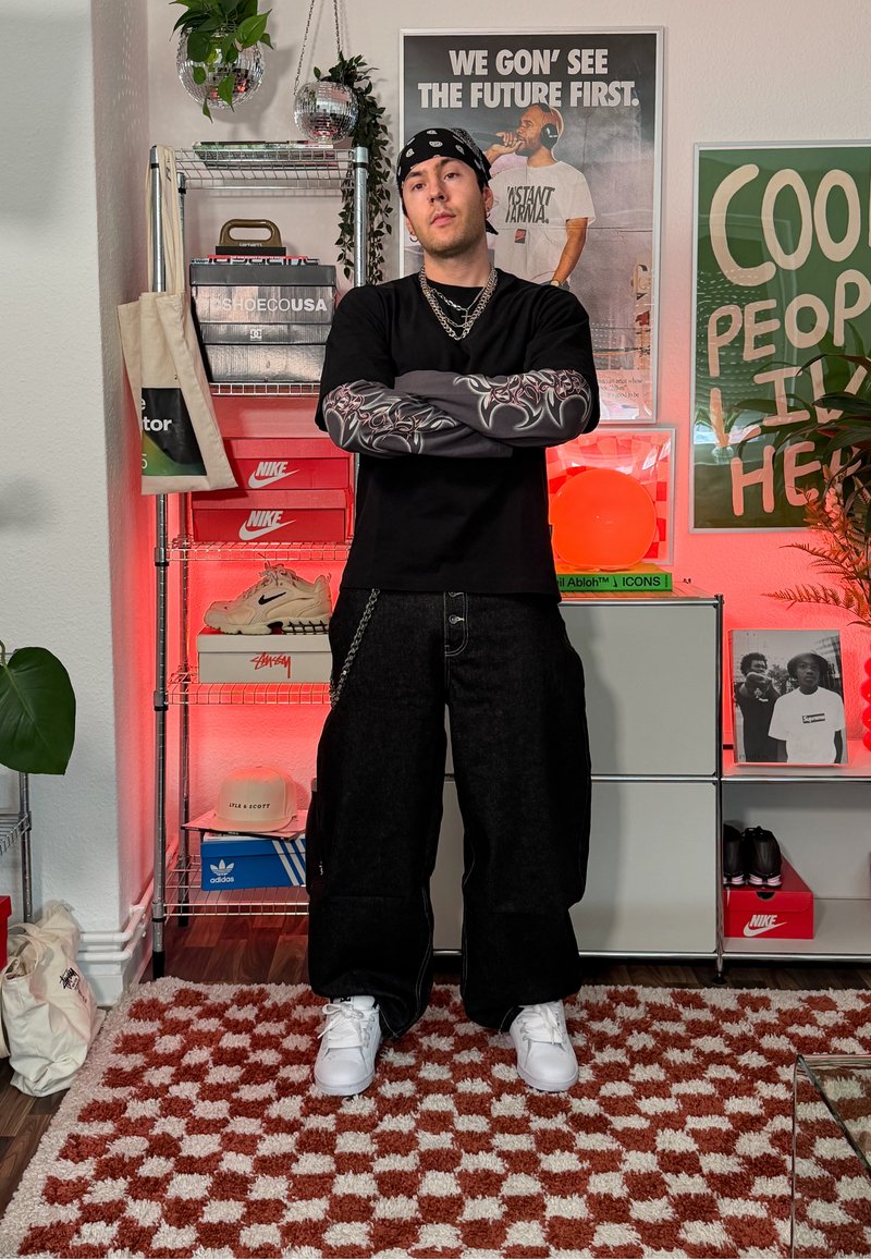 Jeune homme portant un bandana et un collier à chaîne, les bras croisés, devant des boîtes de baskets et des affiches dans une pièce élégante éclairée en rouge.
