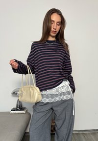 Femme portant une chemise rayée bleu marine avec bordure en dentelle, pantalon gris, collier argenté, tenant un sac à main beige, debout dans une pièce moderne et minimaliste.