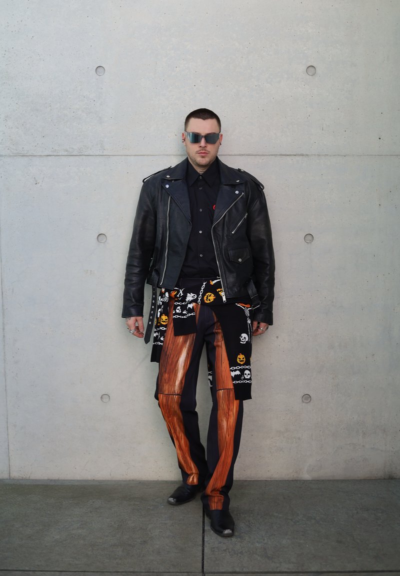 Man wearing black leather jacket, skull and pumpkin patterned sweater tied at waist, wood-patterned pants, and sunglasses against concrete wall.