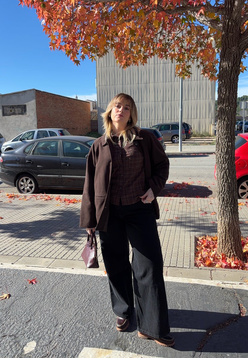 Hnedá nadrozmerná bunda, károvaná košeľa, široké čierne nohavice a hnedé topánky. Drží malú bordovú tašku. Na pozadí jesenné lístie a jasná obloha.